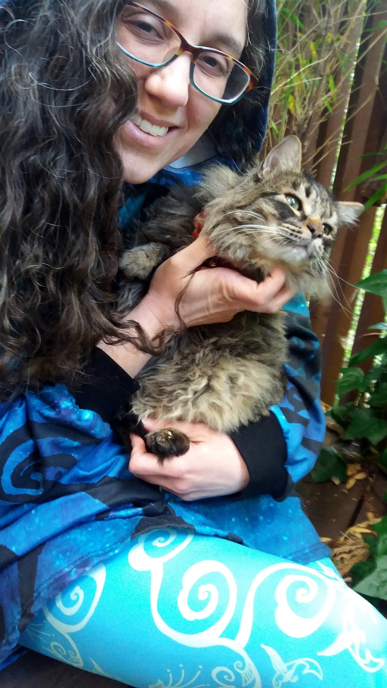 May include: A woman with long brown hair wearing a blue and black patterned hoodie holds a brown tabby cat in her arms. The cat is looking at the camera.