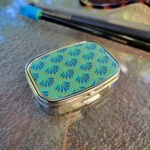 May include: A rectangular, silver-toned pill box with a teal and gold fan-shaped pattern on the lid. The box is closed and sits on a reflective surface. Pens and glasses are in the background.