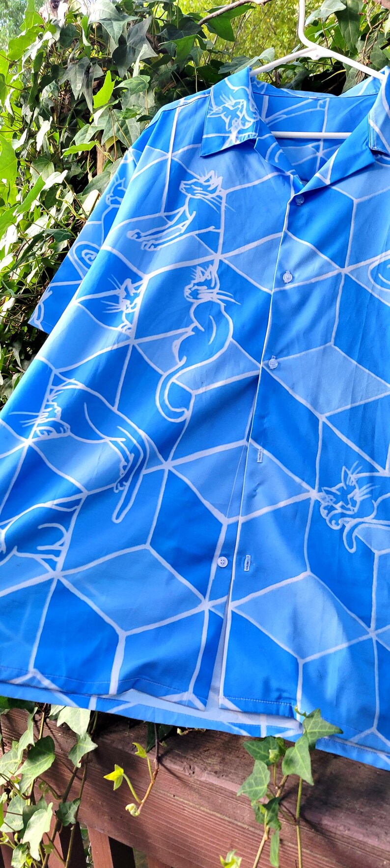 Puede incluir: Camisa azul claro con un dise&ntilde;o geom&eacute;trico blanco e ilustraciones de gatos. La camisa abotonada cuelga de una percha blanca. Fondo de follaje verde.