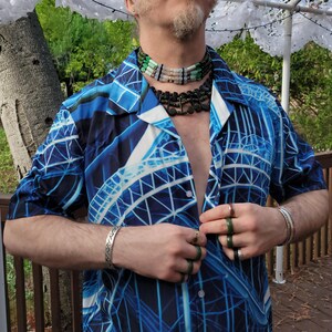 May include: A man wearing a blue and white patterned short-sleeved shirt with a multi-layered beaded necklace and several rings on his fingers.