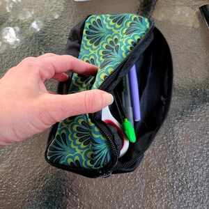 May include: A black pencil case with a teal and yellow floral pattern is partially unzipped, revealing pens, scissors, and markers. The case is held open by a hand, showcasing the interior contents.