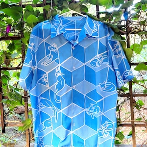 Puede incluir: Camisa azul de manga corta con un patr&oacute;n geom&eacute;trico de cubos azules claros y oscuros. Ilustraciones de gatos blancos est&aacute;n dispersas por toda la camisa. La camisa est&aacute; colgada en una percha blanca.