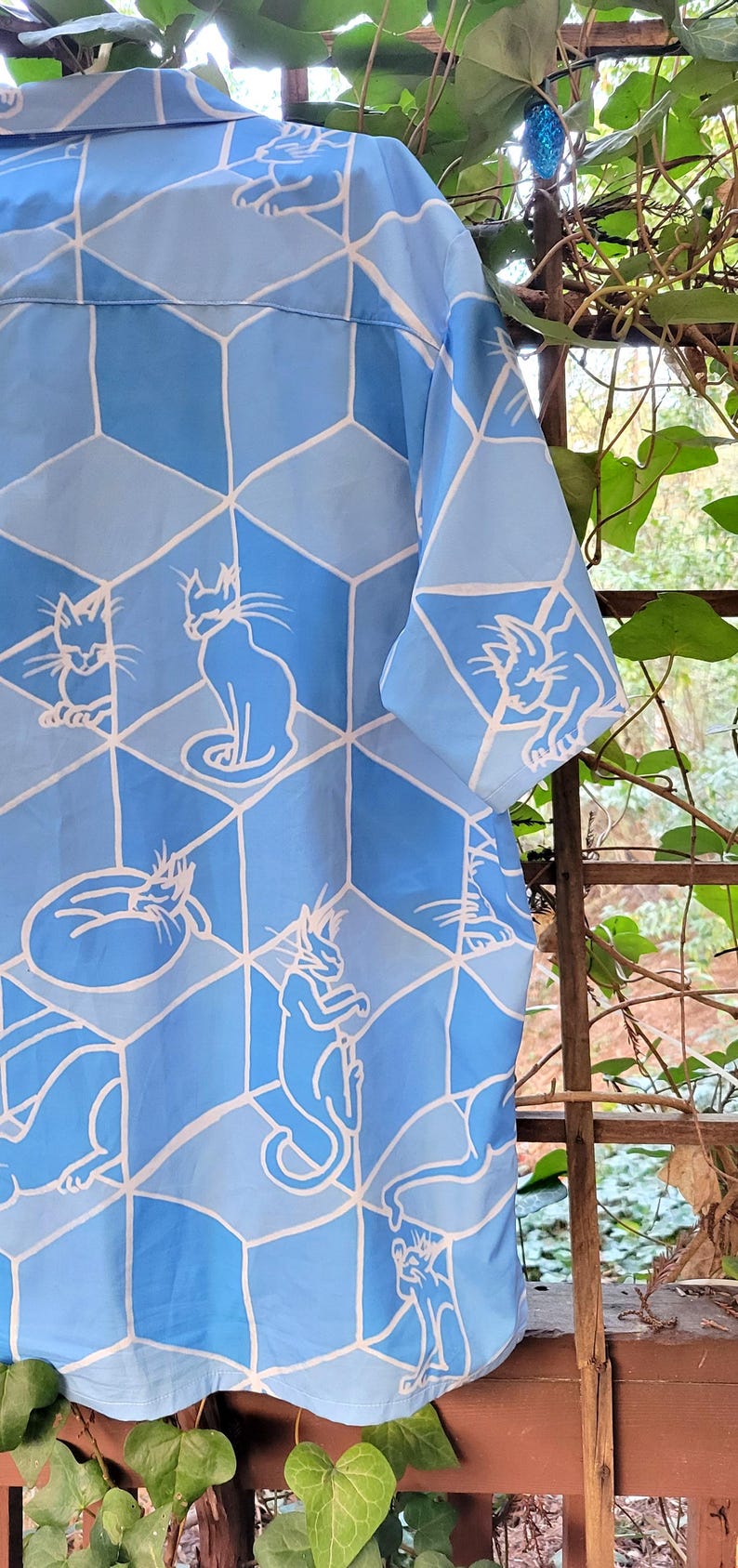 Puede incluir: Camisa azul claro con estampado de gatos. El dise&ntilde;o presenta contornos blancos de gatos dentro de un patr&oacute;n de cubos geom&eacute;tricos en tonos azules. La camisa tiene mangas cortas.