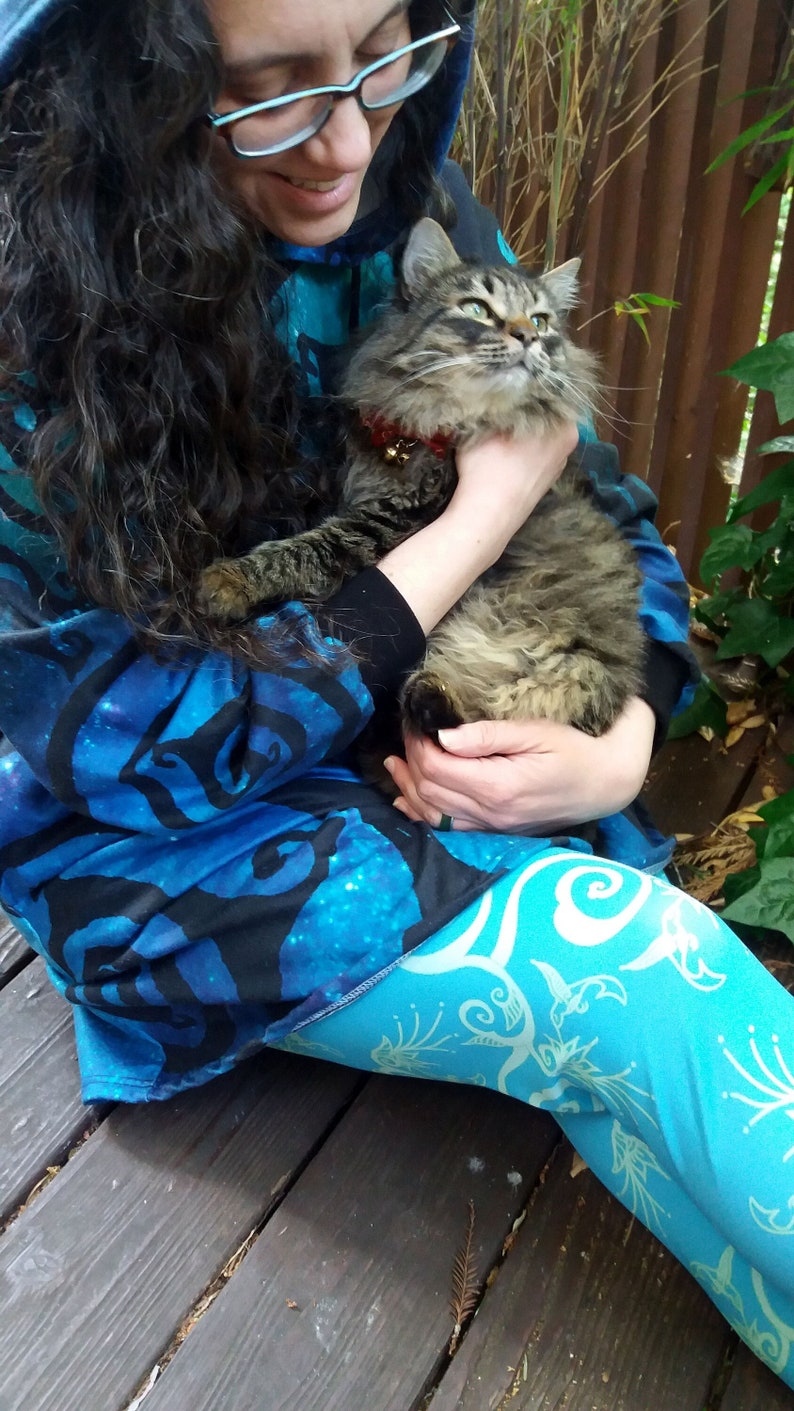 May include: A person wearing a blue and black patterned hoodie and leggings is holding a brown tabby cat. The cat is wearing a gold collar with a bell.