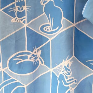 Puede incluir: Tela azul claro con un patr&oacute;n repetido de ilustraciones de gatos blancos dentro de un dise&ntilde;o geom&eacute;trico. Los gatos se representan en varias poses, incluyendo sentados, acurrucados y de pie.