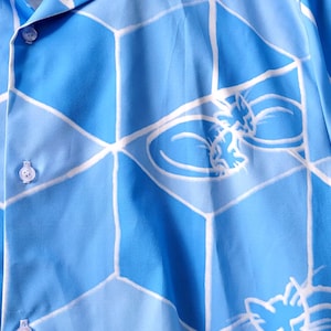 Puede incluir: Camisa azul claro con botones y un patr&oacute;n geom&eacute;trico de l&iacute;neas blancas y formas azules. La camisa presenta un dise&ntilde;o repetido de siluetas de gatos estilizadas. La camisa est&aacute; colgada en una percha blanca.