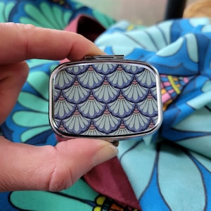 May include: A rectangular, silver-toned pill box with a hinged lid. The top features a repeating pattern of blue and teal fan-shaped designs with pink accents. The box is held against a backdrop of patterned fabric.