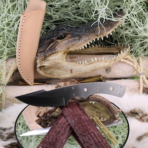 Può includere: Un coltello a lama nera con un fodero in pelle marrone chiaro, sullo sfondo di una testa di alligatore ed elementi decorativi. Il coltello ha un manico scuro ed è esposto su una superficie a specchio.