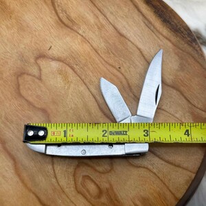 May include: A small, silver pocket knife with three open blades, measured at approximately 3 inches long. The knife is placed on a wooden surface with a yellow measuring tape next to it.