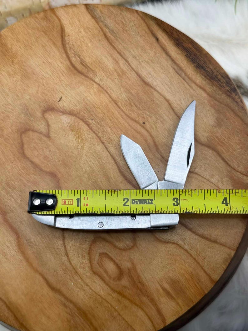 May include: A small, silver pocket knife with three open blades, measured at approximately 3 inches long. The knife is placed on a wooden surface with a yellow measuring tape next to it.