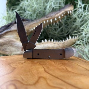 May include: A small, open pocket knife with two blades. The knife has a brown handle and silver-colored metal accents. The blades are open, and the knife is resting on a wooden surface. The background features a blurred alligator head.