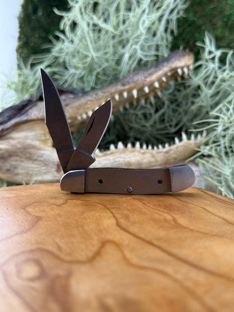 May include: A small, open pocket knife with two blades. The knife has a brown handle and silver-colored metal accents. The blades are open, and the knife is resting on a wooden surface. The background features a blurred alligator head.