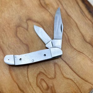 May include: A silver folding pocket knife with two open blades. The knife has a metal handle with rivets. The blades are stainless steel. The knife is resting on a wooden surface.