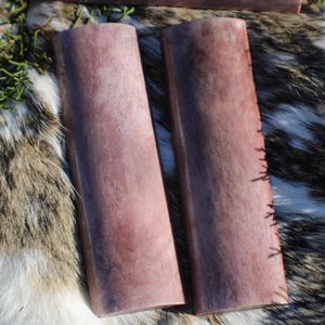 May include: Two purple and white bone knife scales, with a natural wood grain pattern. The scales are about 4 inches long and 1 inch wide.