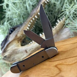 May include: A small, open pocket knife with a silver blade and a brown handle. The knife is partially open, revealing the sharp blade. The background features a wooden surface and a blurred alligator head.