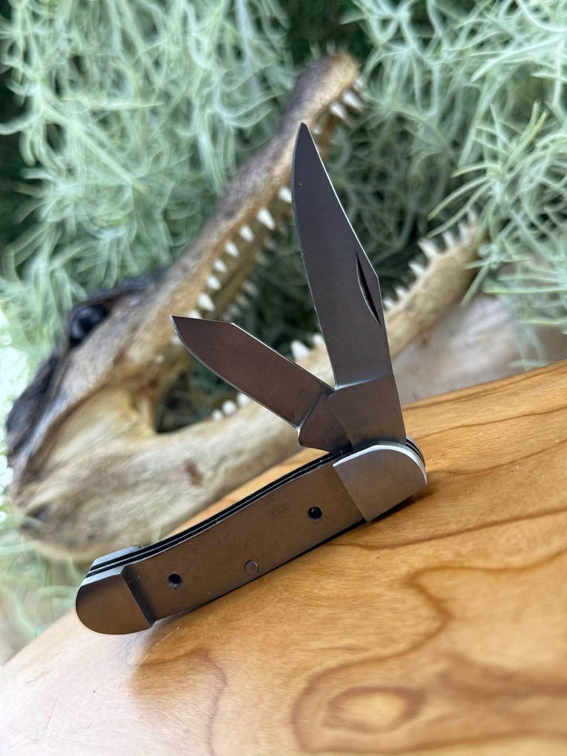 May include: A small, open pocket knife with a silver blade and a brown handle. The knife is partially open, revealing the sharp blade. The background features a wooden surface and a blurred alligator head.