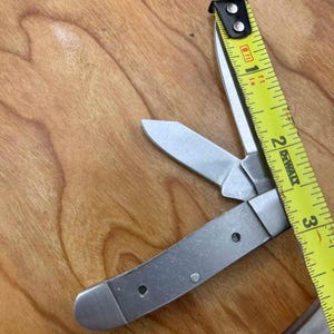May include: A silver folding pocket knife with two open blades and a yellow tape measure. The tape measure shows measurements in inches. The knife is on a wooden surface.