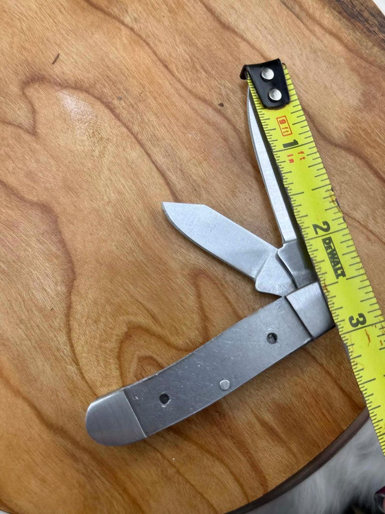 May include: A silver folding pocket knife with two open blades and a yellow tape measure. The tape measure shows measurements in inches. The knife is on a wooden surface.