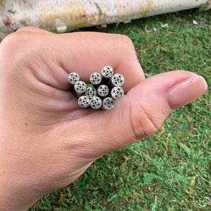 May include: A hand holding a bundle of small, black metal tubes with a patterned design on the ends. The ends of the tubes have a gold and black dot pattern. The background includes green moss and a piece of wood.
