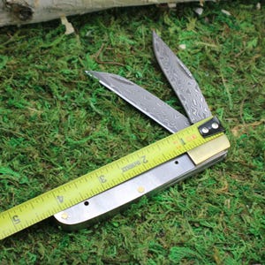 May include: A partially open folding knife with a patterned blade, measured with a yellow tape measure. The knife is on a bed of green moss. The tape measure shows approximately 3 inches.