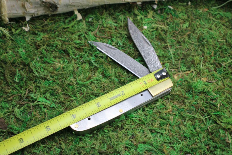 May include: A partially open folding knife with a patterned blade, measured with a yellow tape measure. The knife is on a bed of green moss. The tape measure shows approximately 3 inches.