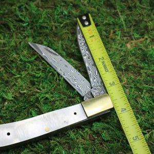 May include: A partially open folding knife with a patterned blade and brass accents. A yellow measuring tape is positioned diagonally across the knife, showing measurements in inches. The knife rests on a bed of green moss.