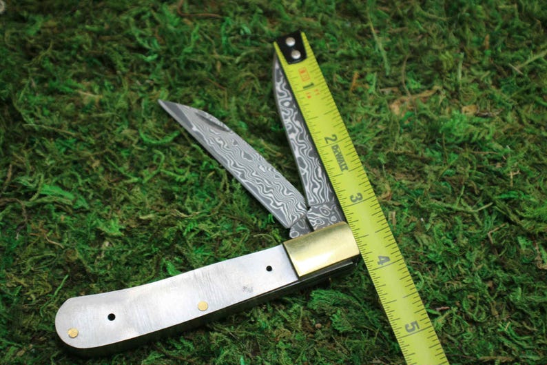 May include: A partially open folding knife with a patterned blade and brass accents. A yellow measuring tape is positioned diagonally across the knife, showing measurements in inches. The knife rests on a bed of green moss.