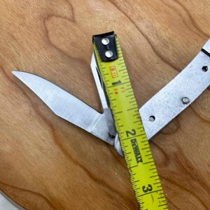 May include: A multi-tool knife with a partially open blade, next to a yellow and black measuring tape. The tape reads 2 inches. The knife and tape are on a wooden surface.