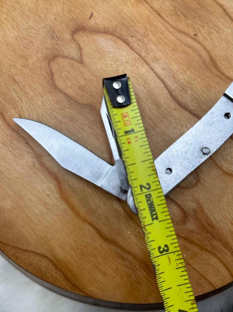 May include: A multi-tool knife with a partially open blade, next to a yellow and black measuring tape. The tape reads 2 inches. The knife and tape are on a wooden surface.