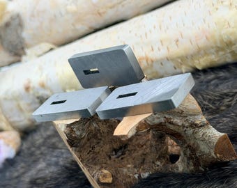 May include: Three rectangular, silver-colored metal cufflinks with a rectangular cutout in the center. The cufflinks are displayed on a piece of wood, with a backdrop of birch logs and a dark, textured surface.