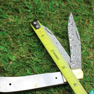 May include: A folding pocket knife with a patterned blade is open, next to a yellow tape measure. The knife has a silver handle with brass rivets and a cream-colored bolster. The tape measure reads 3 inches.