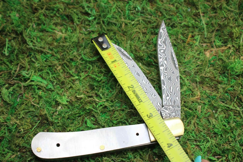 May include: A folding pocket knife with a patterned blade is open, next to a yellow tape measure. The knife has a silver handle with brass rivets and a cream-colored bolster. The tape measure reads 3 inches.