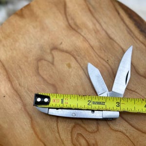 May include: A small, silver folding knife with two open blades, next to a yellow tape measure. The tape measure reads approximately 3 inches. The knife and tape measure are on a wooden surface.