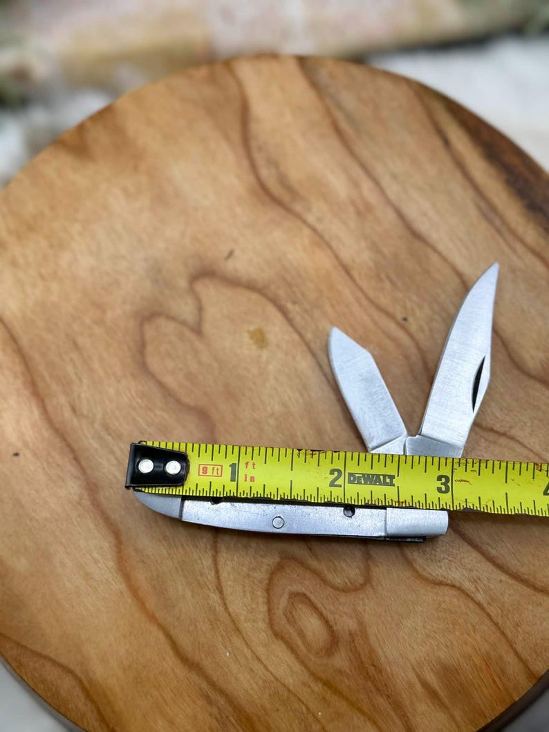 May include: A small, silver folding knife with two open blades, next to a yellow tape measure. The tape measure reads approximately 3 inches. The knife and tape measure are on a wooden surface.