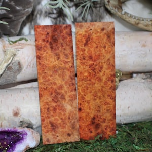 May include: Two rectangular wooden pieces with a warm, reddish-brown burl grain pattern. They are set against birch logs, greenery, and a geode. The wood pieces are likely for crafting or woodworking, possibly for a project.