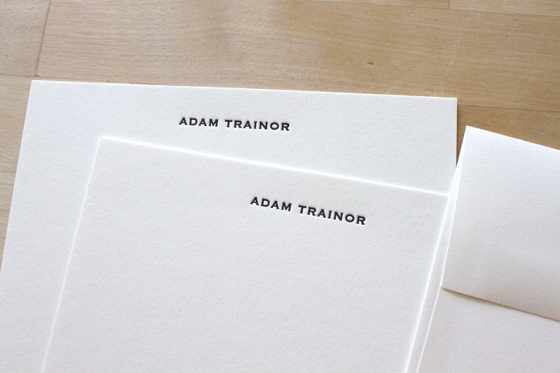 Two white cards with the text 'ADAM TRAINOR' letterpress printed in black on each card. The cards are stacked on top of each other.