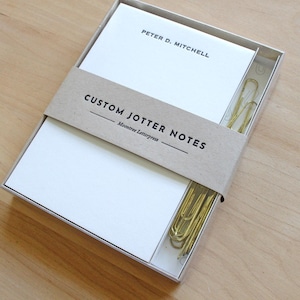 May include: A white notepad set in a clear box, with gold paper clips. The notepad is wrapped with a brown band that reads "CUSTOM JOTTER NOTES". The top notepad has the name "PETER D. MITCHELL" printed on it.