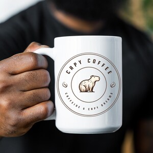 May include: White ceramic coffee mug with a brown logo that says "Capy Coffee" and features a cartoon capybara. The logo also includes the text "Caffeine & Capy Dreams".