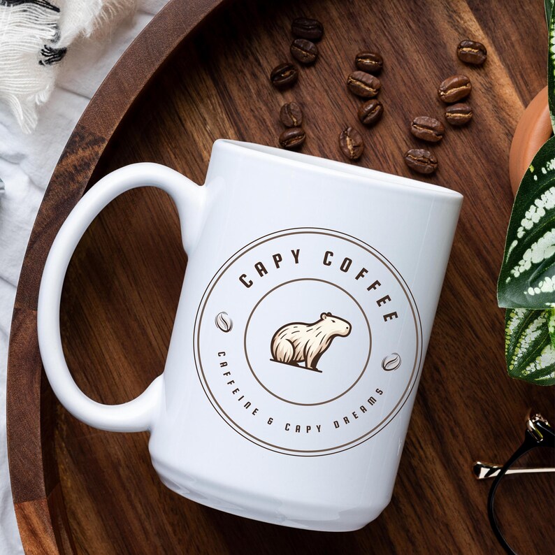 May include: White ceramic coffee mug with a brown circle design featuring a cartoon capybara and the text "Capy Coffee Caffeine & Capy Dreams".