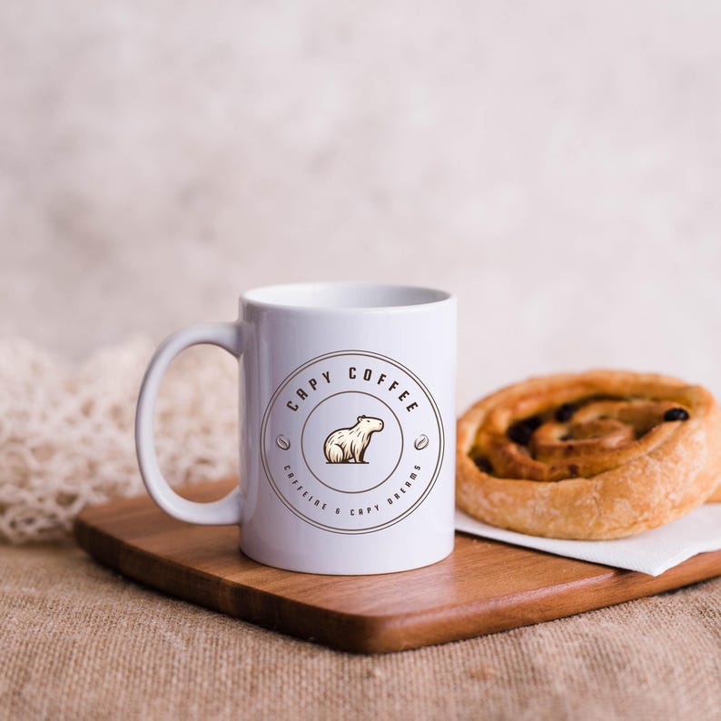 May include: White ceramic coffee mug with a brown logo that says "Capy Coffee" and features a cartoon capybara. The mug is on a wooden cutting board with a pastry next to it.