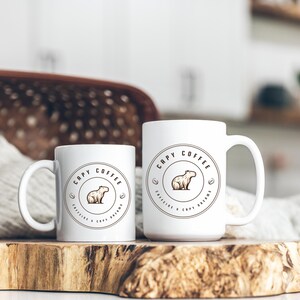 May include: Two white ceramic mugs with a brown capybara logo. The logo says "Capy Coffee Caffeine & Capy Dreams".