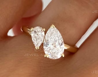 May include: A gold ring featuring two pear-cut clear stones set in a bypass style. The larger pear-cut stone is positioned next to a smaller pear-cut stone on a polished gold band. The text JEWEL NESTS is visible in the image.