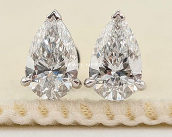 Könnte beinhalten: Ein Paar tropfenförmige Diamantohrstecker. Die Diamanten sind in einem silberfarbenen Metall gefasst und werden auf einer cremefarbenen Oberfläche präsentiert. Klassischer Schmuck.