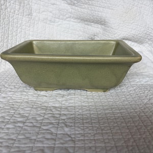 May include: A rectangular ceramic bonsai pot with a muted sage green glaze. The vessel features a wide, flat rim, slightly tapered sides, and a shallow basin. The surface has a subtle, speckled texture throughout the matte finish.