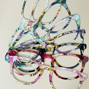 May include: A stack of eyeglasses with colorful, abstract patterns. The frames display a variety of hues, including pink, purple, blue, and yellow, creating a vibrant and artistic design. The glasses are stacked on top of each other.