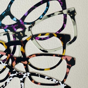 Puede incluir: Una pila de cinco pares de gafas con diferentes marcos estampados. Los marcos son de varios colores, incluyendo negro, blanco, azul, rosa y amarillo.