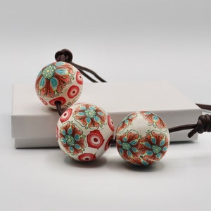 May include: A necklace with three large, round, white beads with a red, blue, and green floral pattern. The beads are strung on a brown leather cord.