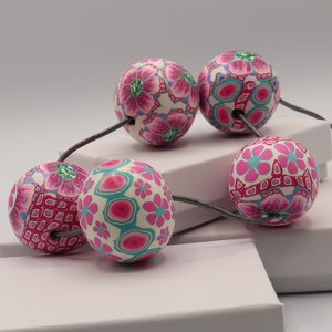 May include: A necklace with five large, round beads. The beads are made of polymer clay and have a pink, white, and teal floral pattern. The necklace is strung on a gray cord.