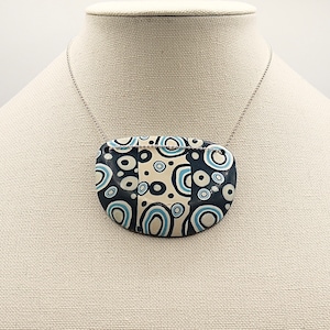 May include: A necklace with a large, oval pendant featuring a geometric pattern of black, white, and blue circles. The pendant is made of polymer clay and hangs from a silver chain.