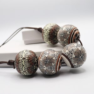 May include: A necklace made of large, round, brown and white beads with intricate floral and geometric patterns. The beads are strung on a brown cord.